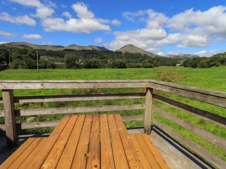 A wooden picnic table on a wooden deck overlooking green fields and hills under a blue sky at Ellis Howe in Torver