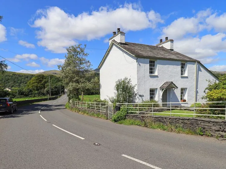 A white house next to a curved road with trees and hills in the background at Ellis Howe in Torver